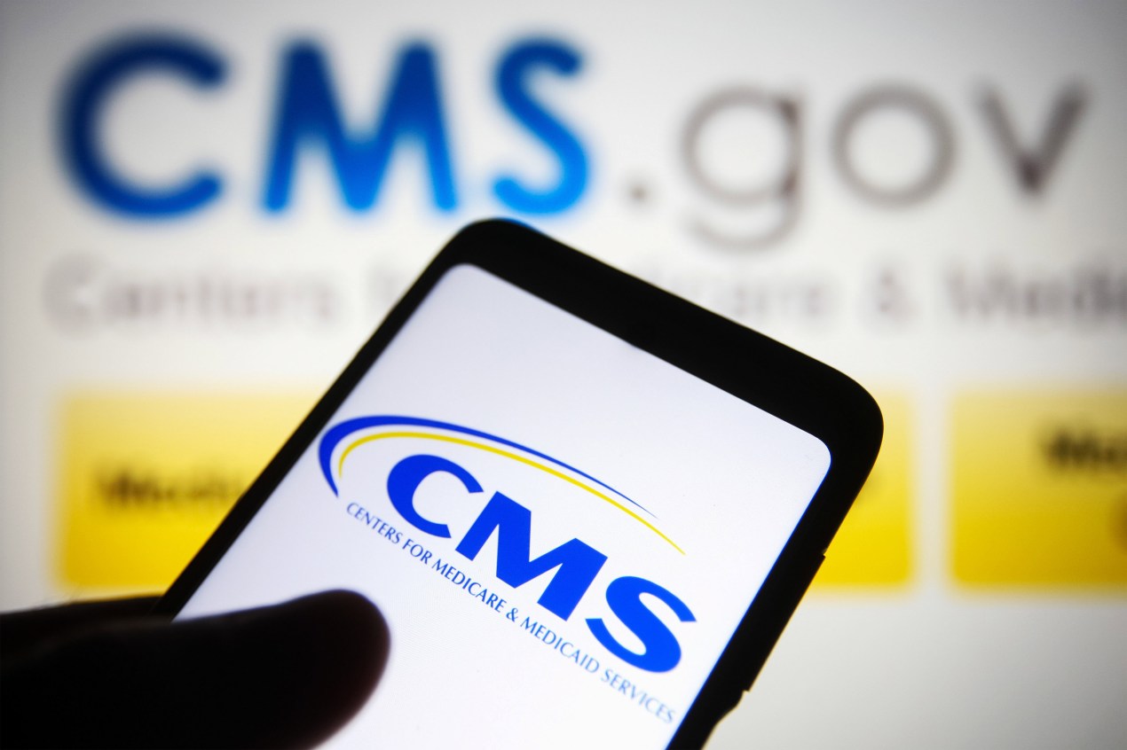 A photo shows the CMS logo on a phone screen held in front of a computer monitor that shows the CMS logo.
