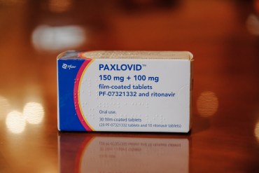 A box of 'Paxlovid' is in the center of the image. The box is white with blue text, with a thick a blue and red stripe on its left.