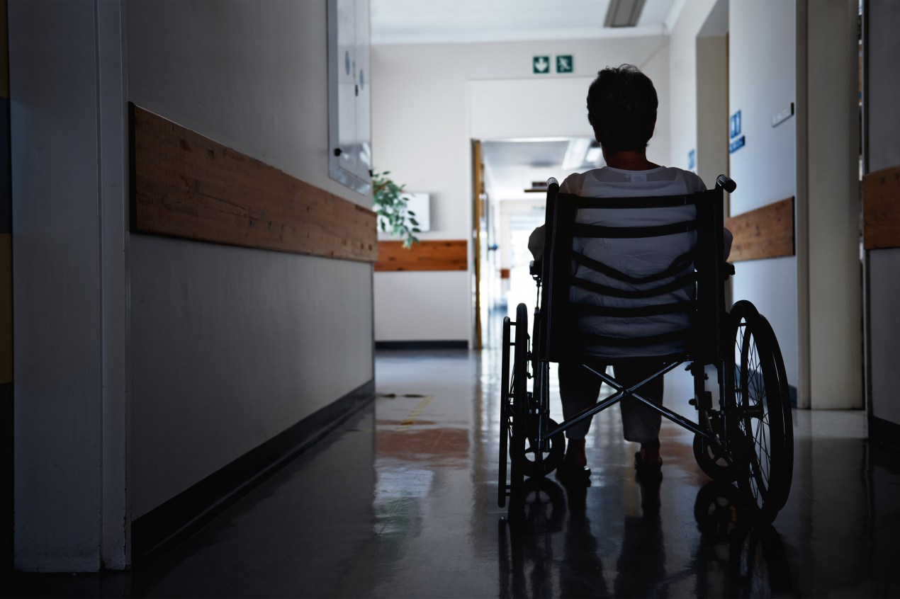 A photo shows a senior woman sitting in a wheelchair in a nursing home corridor.