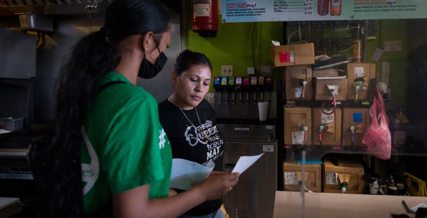 Melissa Lopez explains the covid testing process to a taco shop employee. They are in the back of the restaurant, and storage items are visible on the shelves behind them.