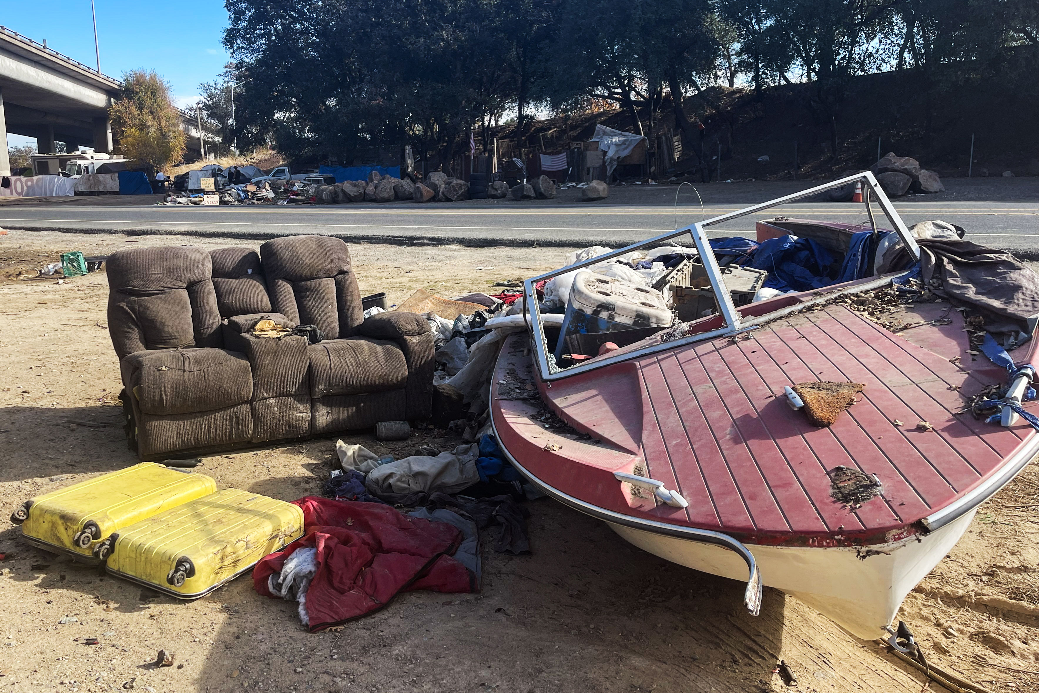 A photo shows a homeless encampment on the side of a road. A couch and boat are on the side of the road and filled with discarded items.