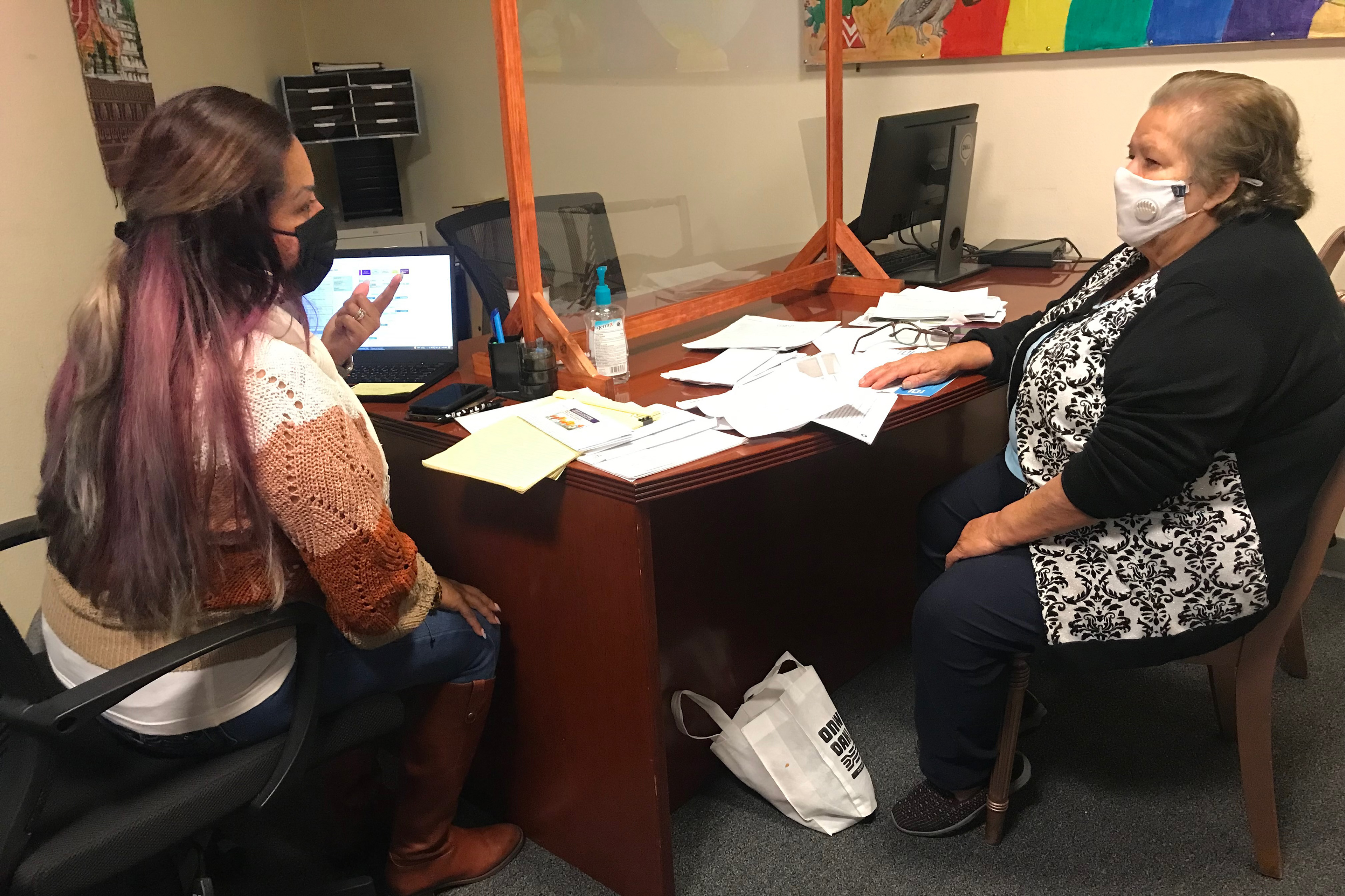A health educator speaks with a client in an office. They are sitting tangentially beside each other at a desk covered in paperwork. 