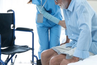 A photo shows an older man being helped into a wheelchair by a nurse.