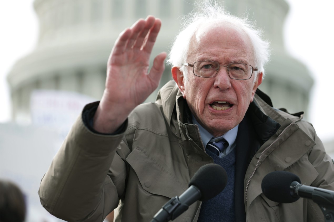 A photo shows Senator Bernie Sanders speaking into a microphone in front of the U.S. Capitol.