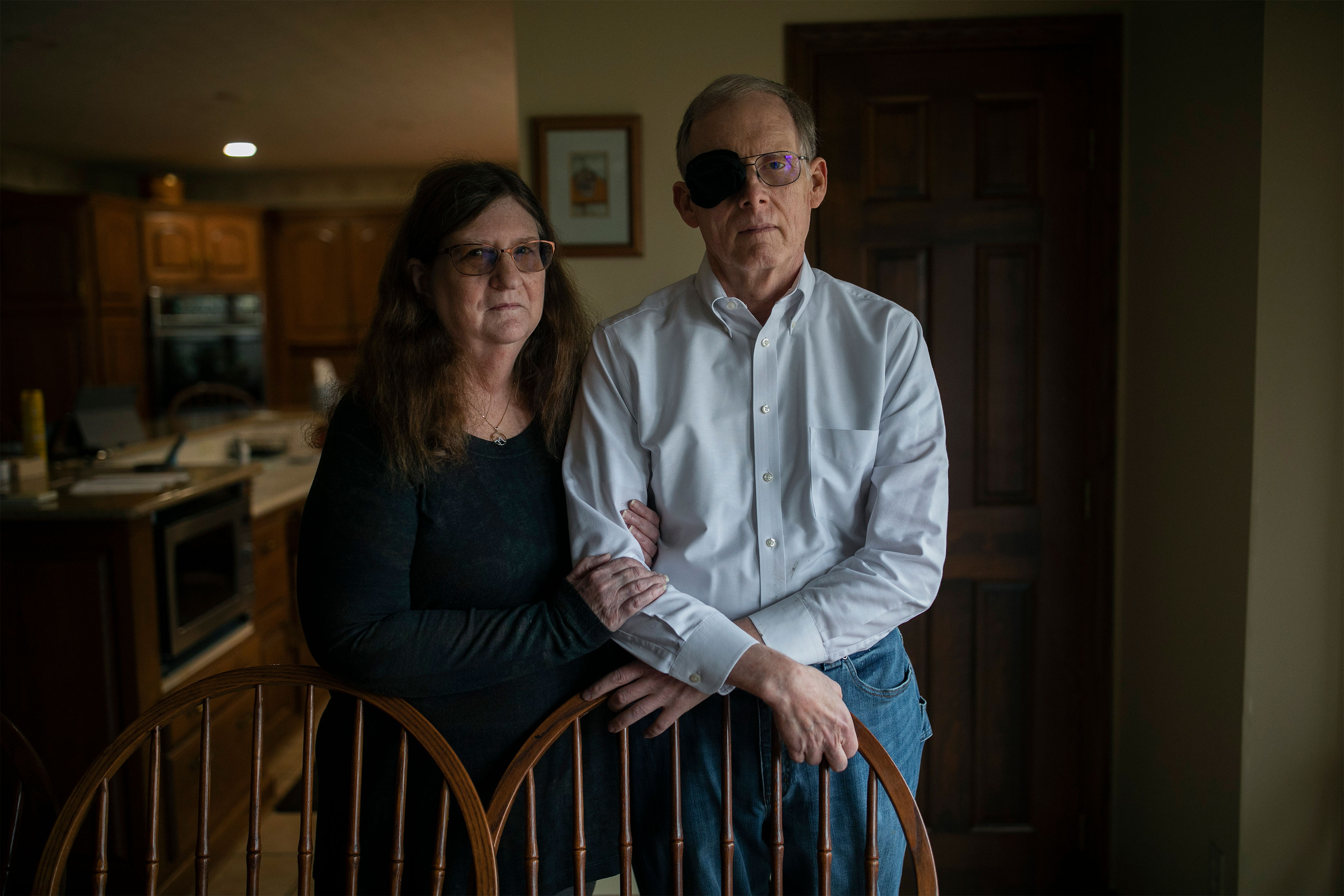 A woman and a man stand behind two dining room chairs. They are both wearing glasses and the man has a black cloth covering his right eye over his glasses. She is gently holding his right arm with both hands.