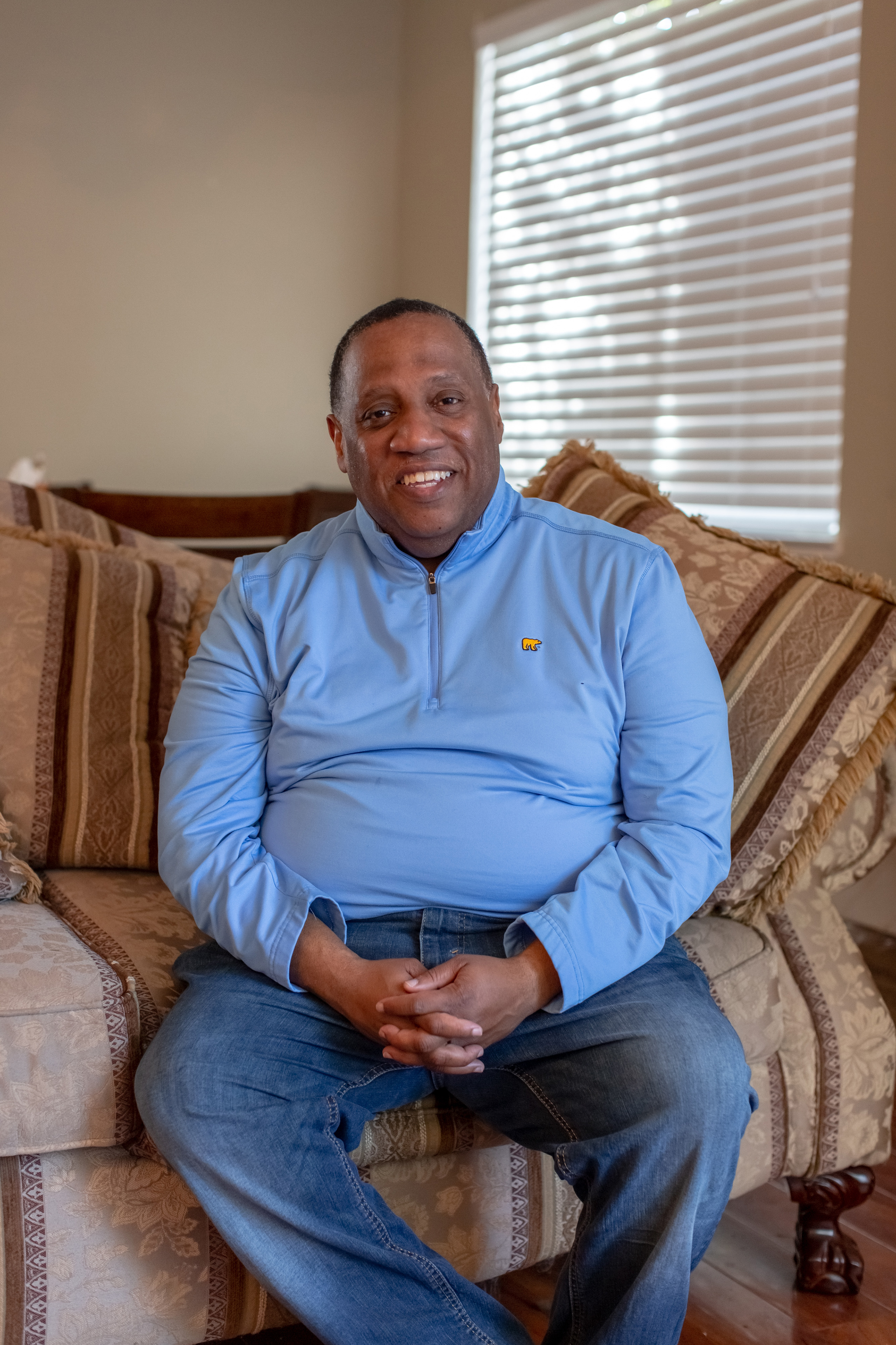 Ovester Armstrong Jr. sits on a couch in his home and holds his hands in his lap. He is looking directly towards the camera and smiling broadly.