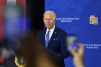 A photo shows President Joe Biden speaking at a podium while blurred hands hold up cellphones in the foreground.