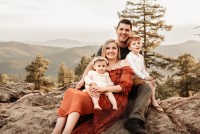 Brittany Tesso is pictured with her family in a nicely staged professional photograph. Her youngest child sits in her lap, while her husband, who is seated beside her, holds their older son. They are outdoors in a mountainous area.
