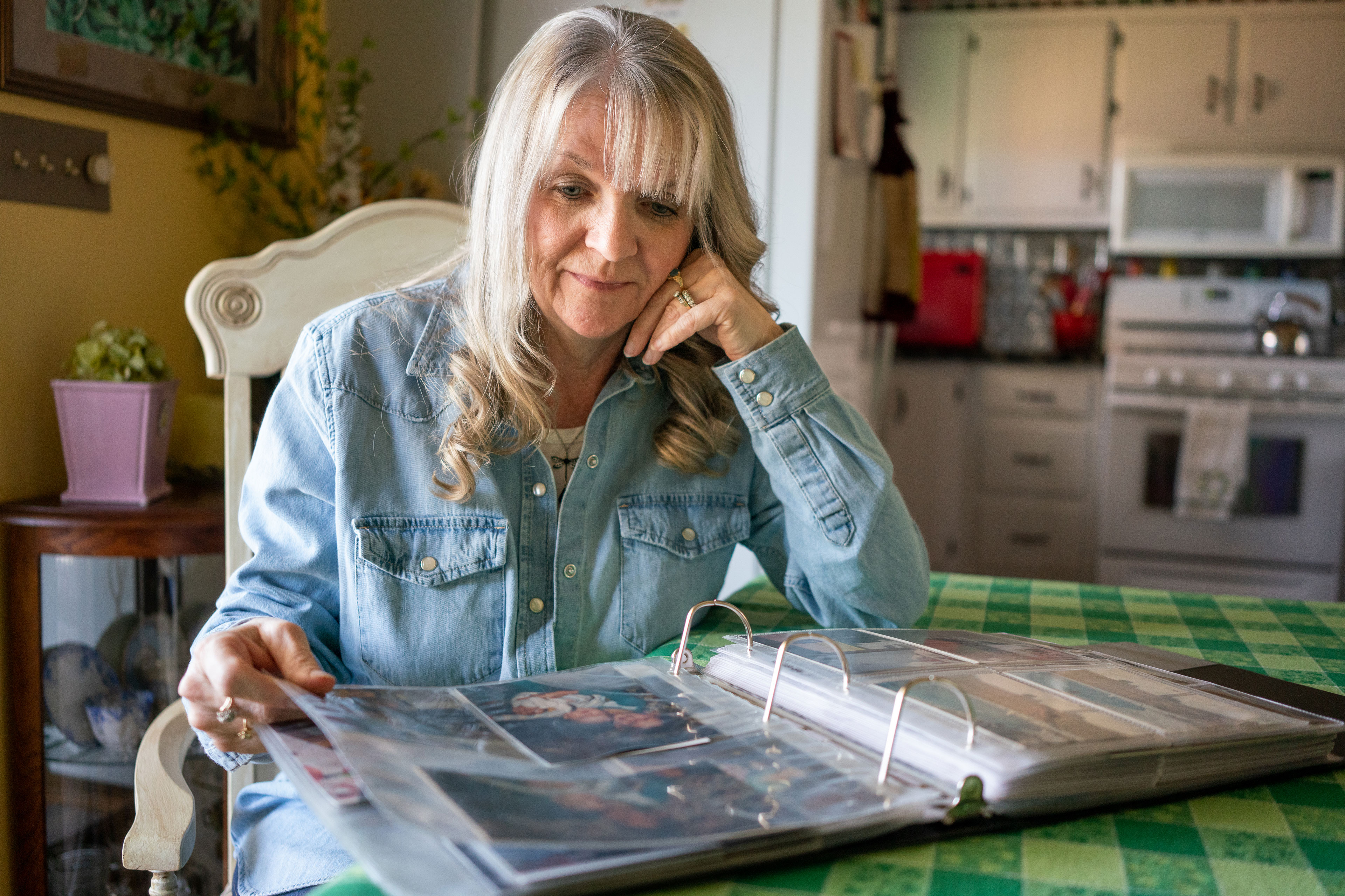 A woman sits at a kitchen table, the stove in the background, her gaze peering down at a photo album. She has light hair and is wearing a light blue denim shirt. The photos in the album appear to be of young children.
