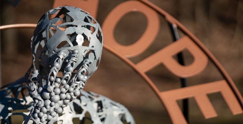 A close-up image of a metal statue of a person leaning their head on their hand. Slightly out of focus behind the figure is the word "Hope" in copper -- another part of the statue.