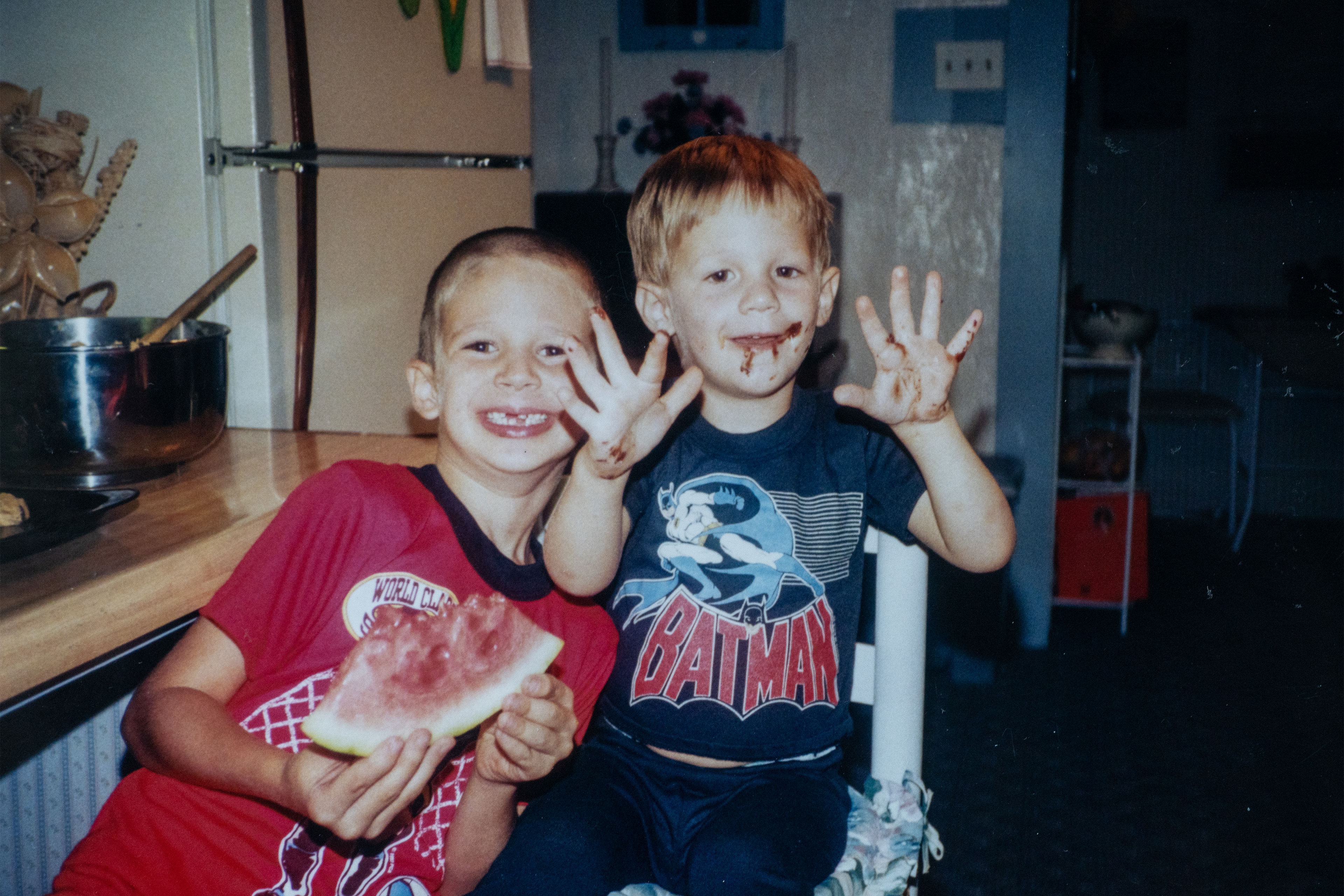 Two children, both boys, smile at the camera. One boy, in a red tshirt, is eating a slice of watermelon and has several teeth missing. The other, in a navy Batman tshirt, holds up his hands which, like his face, are covered in chocolate.