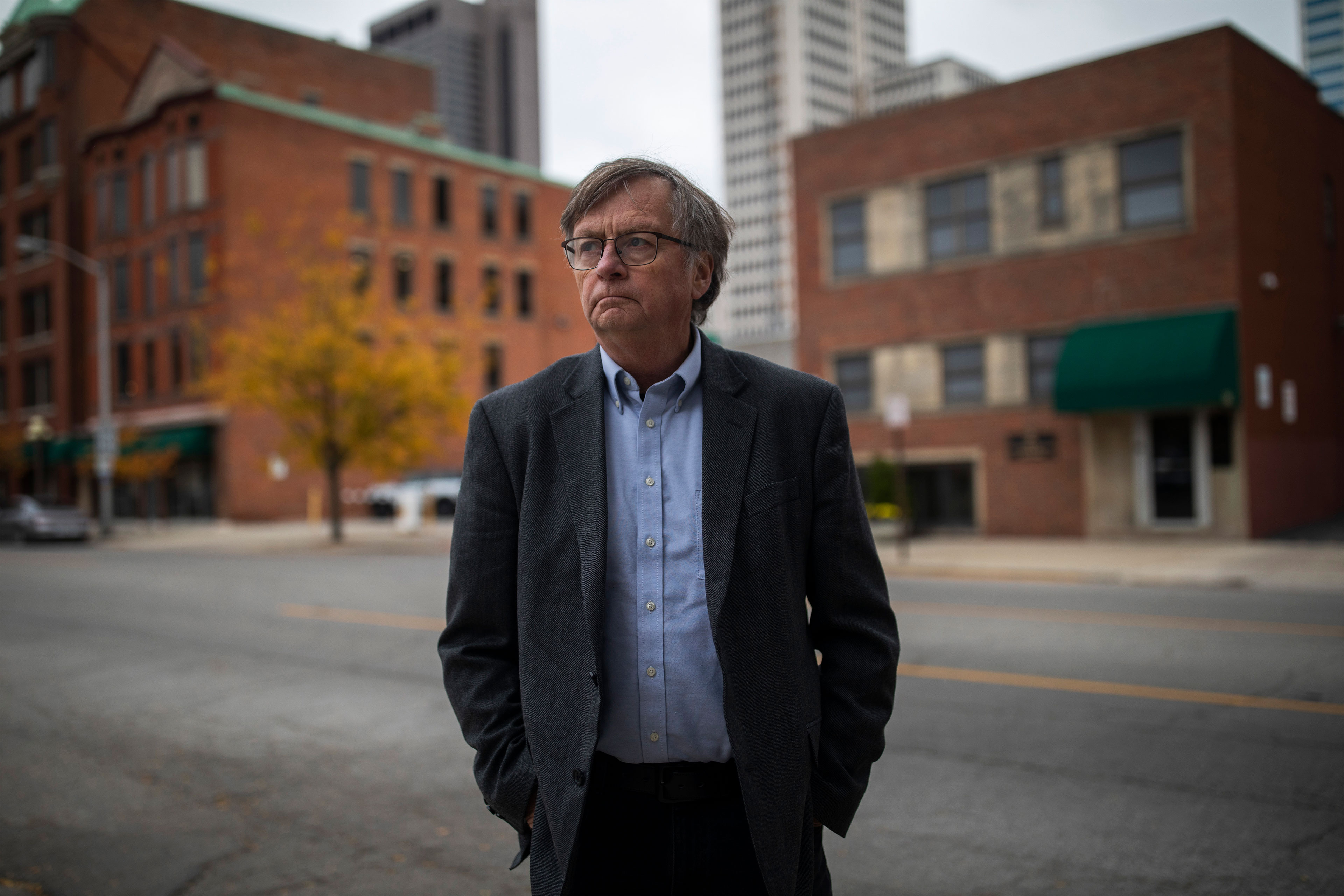 A middle-aged man in a dark grey suit with a light blue buttoned shirt stands on a city street looking to the left. He has a serious expression and wears glasses.