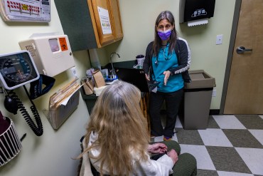 One woman in a mask facing the camera speaks to another woman facing away from the camera in a clinical office.