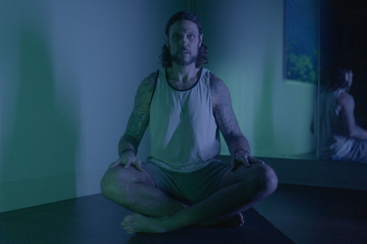 A photo of Riley Cote sitting in a yoga pose in a room softly lit by blue light.