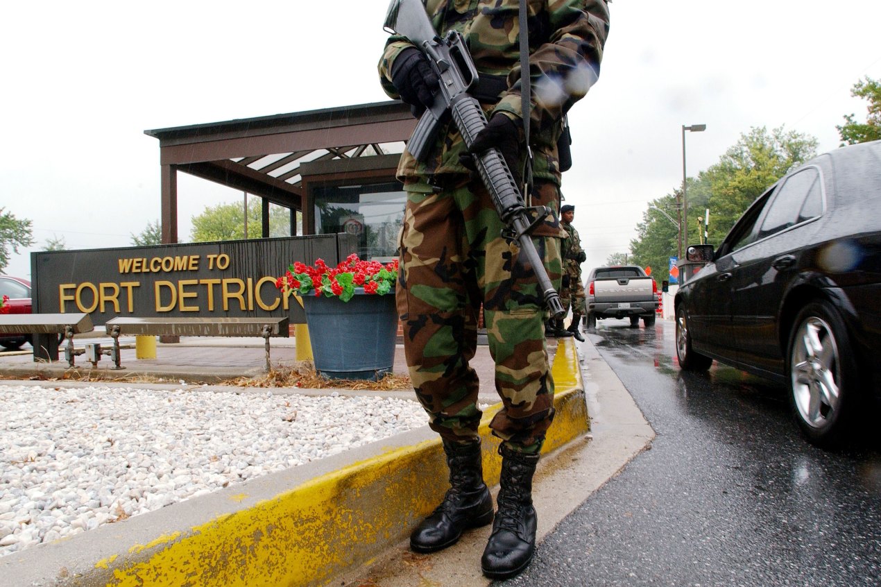 A person wearing military fatigues and holding a gun stands centrally in the foreground. Behind them is a sign that reads "Welcome to Fort Detrick" and cars driving past a guard house.