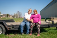 A photo of an older woman with her adult daughter sitting for a portrait outside by a barn.