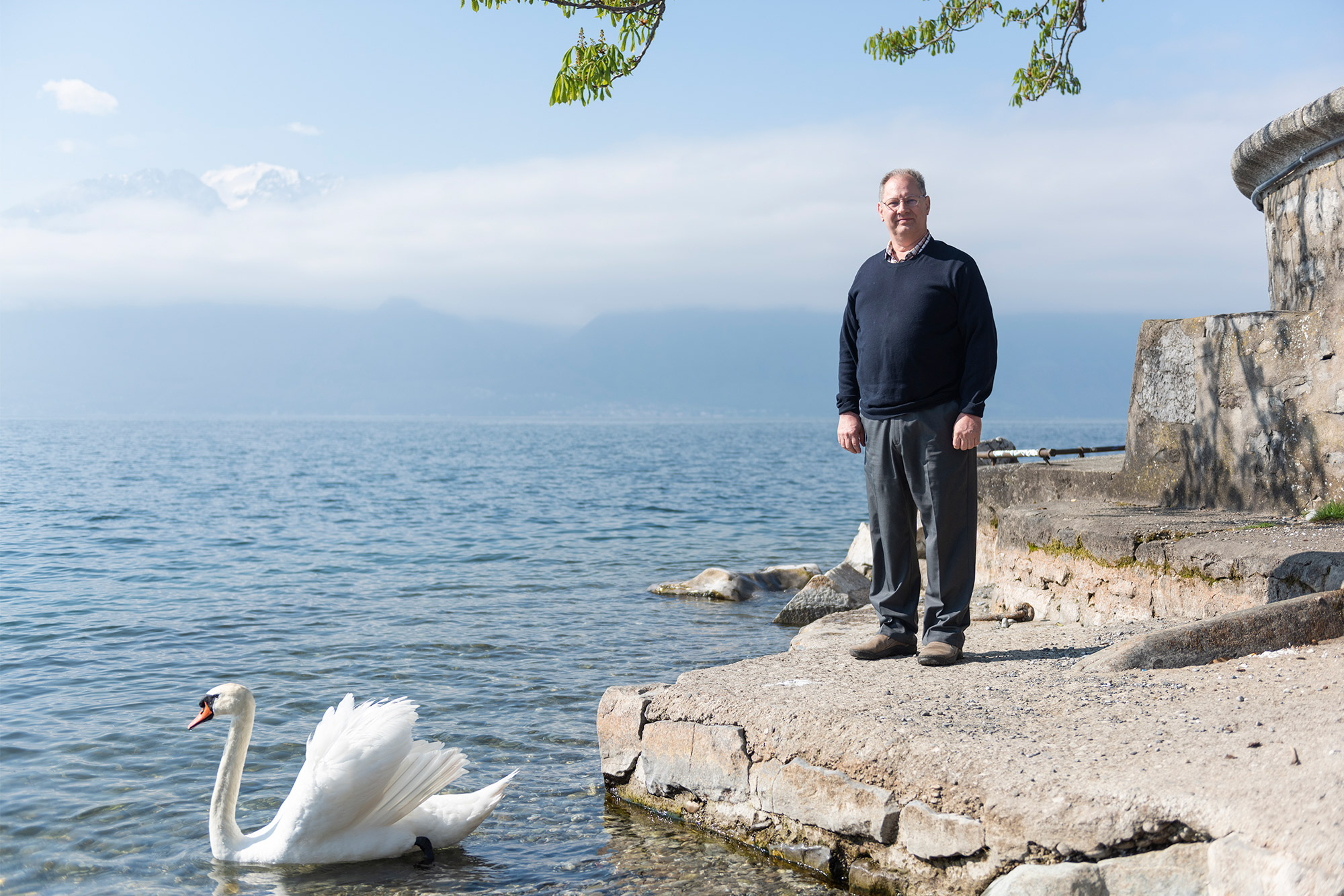 A photo of Jay Comfort standing for a photo by the water as a swan swims nearby.