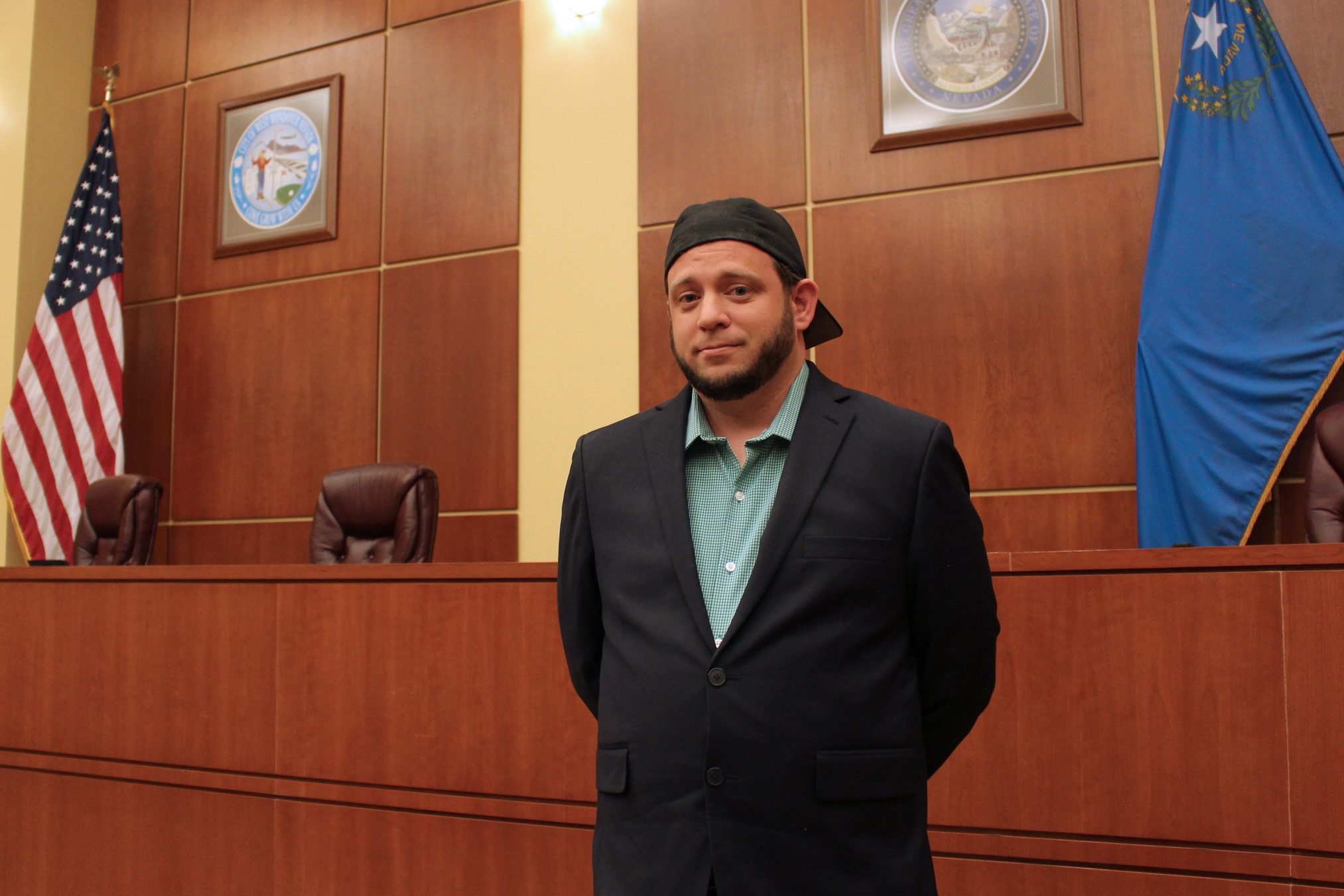 A photo of a man standing for a photo indoors by the Nevada flag and the American flag.