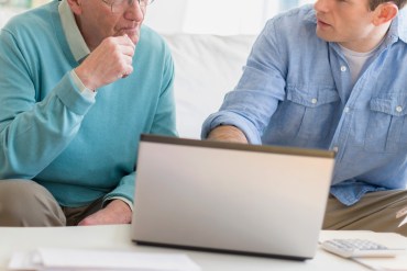 A photo of an older man receiving computer help from a younger man.