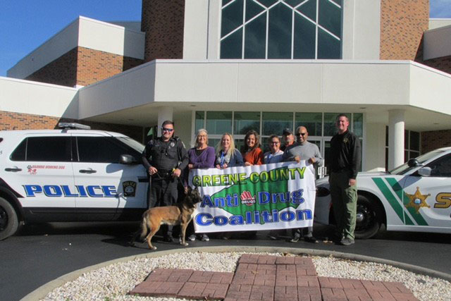 Nine people and a dog stand in front of a building and two police vehicles. Some of the people hold a banner that reads 'Greene County Anti-Drug Coalition'.