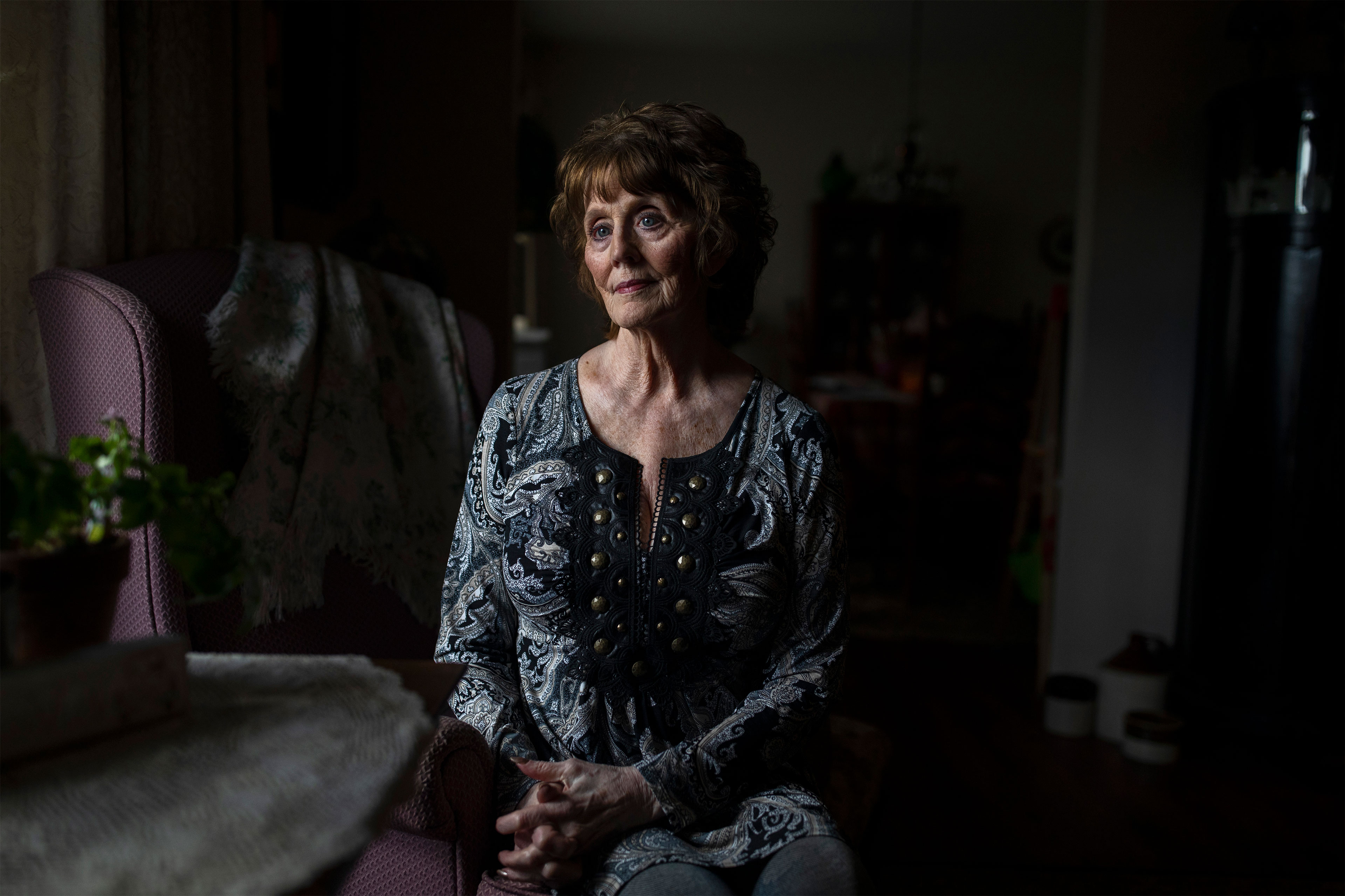 An older woman sits in a darkened room and is illuminated by light through a window.