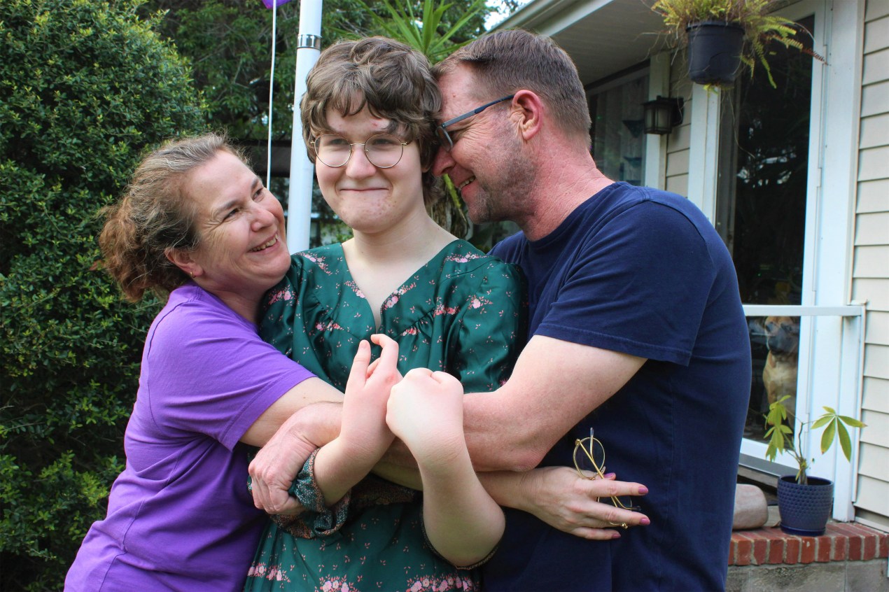 A photo of a teenage transgender girl being hugged by her mother and father outside.