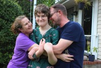 A photo of a teenage transgender girl being hugged by her mother and father outside.