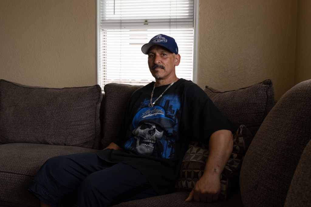 A man with facial hair wearing a baseball cap and dark tshirt sits on a couch facing the camera.