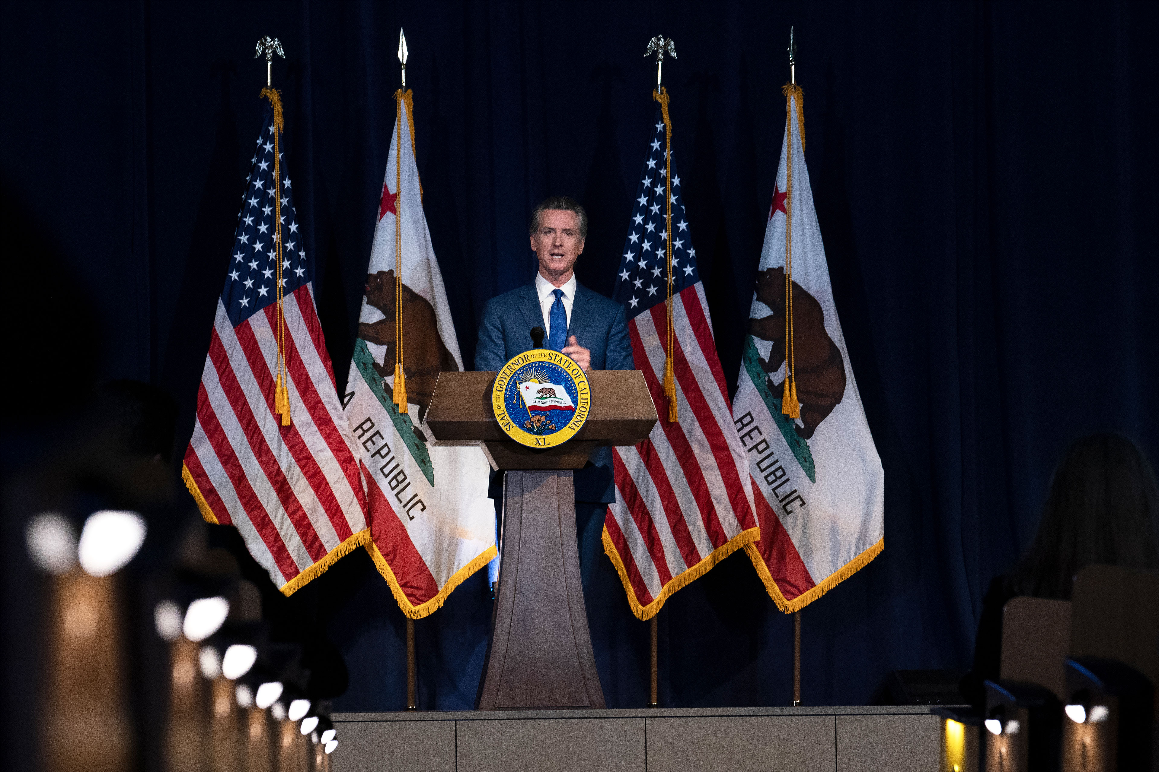Gavino Newsomo nuotrauka, pristatanti savo peržiūrėtą biudžetą už podiumo ir apsupta Amerikos bei Kalifornijos vėliavų.