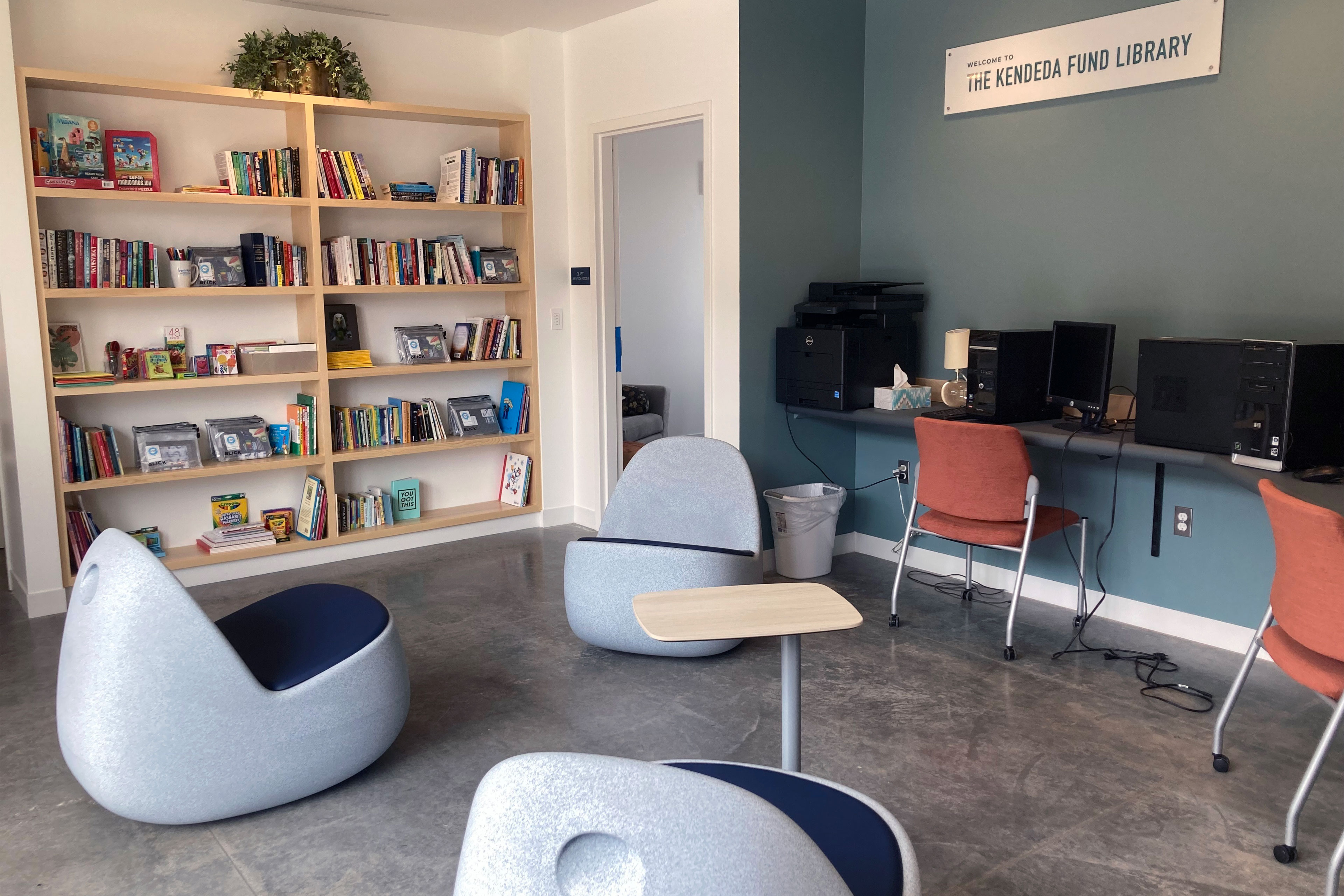 A desk along one wall of the room has computers and printers. There is a bookshelf along another wall and lounge chairs in the middle of the room.