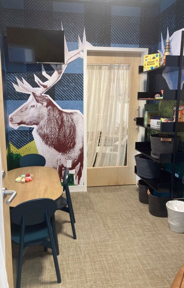 There are child-sized table and chairs and shelves with games in this small room. There is a tv mounted on the wall and an large image of an elk is affixed to the the wall.
