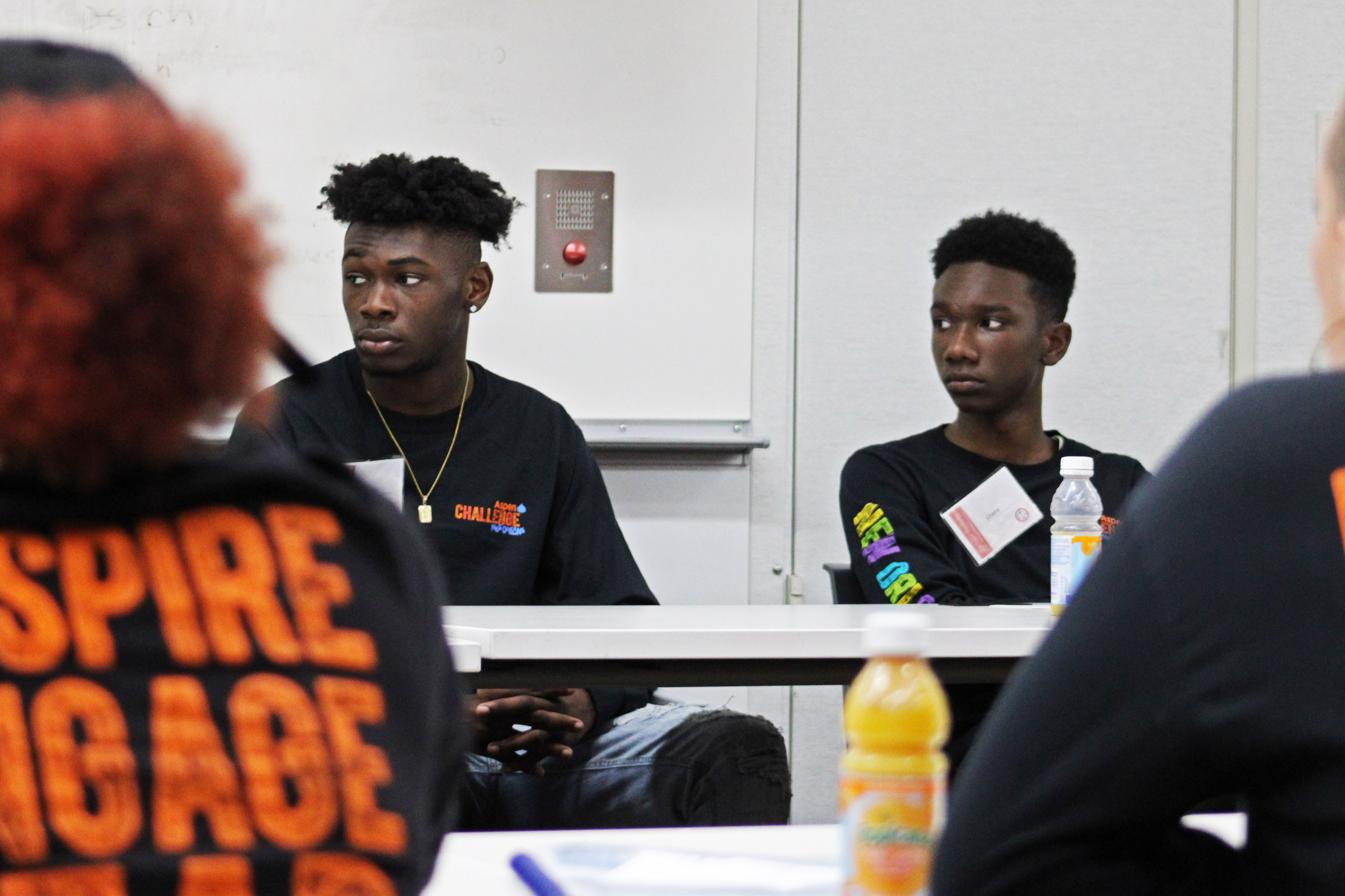 A photo of two Black high schoolers sitting at a table listening to someone speak.