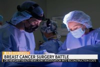 A still from a video of medical workers in surgical gowns and masks. Text on the screen reads, "Breast cancer surgery battle. CBS / KFF Health News investigation into reconstruction costs.