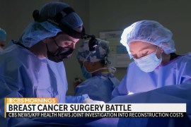 A still from a video of medical workers in surgical gowns and masks. Text on the screen reads, "Breast cancer surgery battle. CBS / KFF Health News investigation into reconstruction costs.