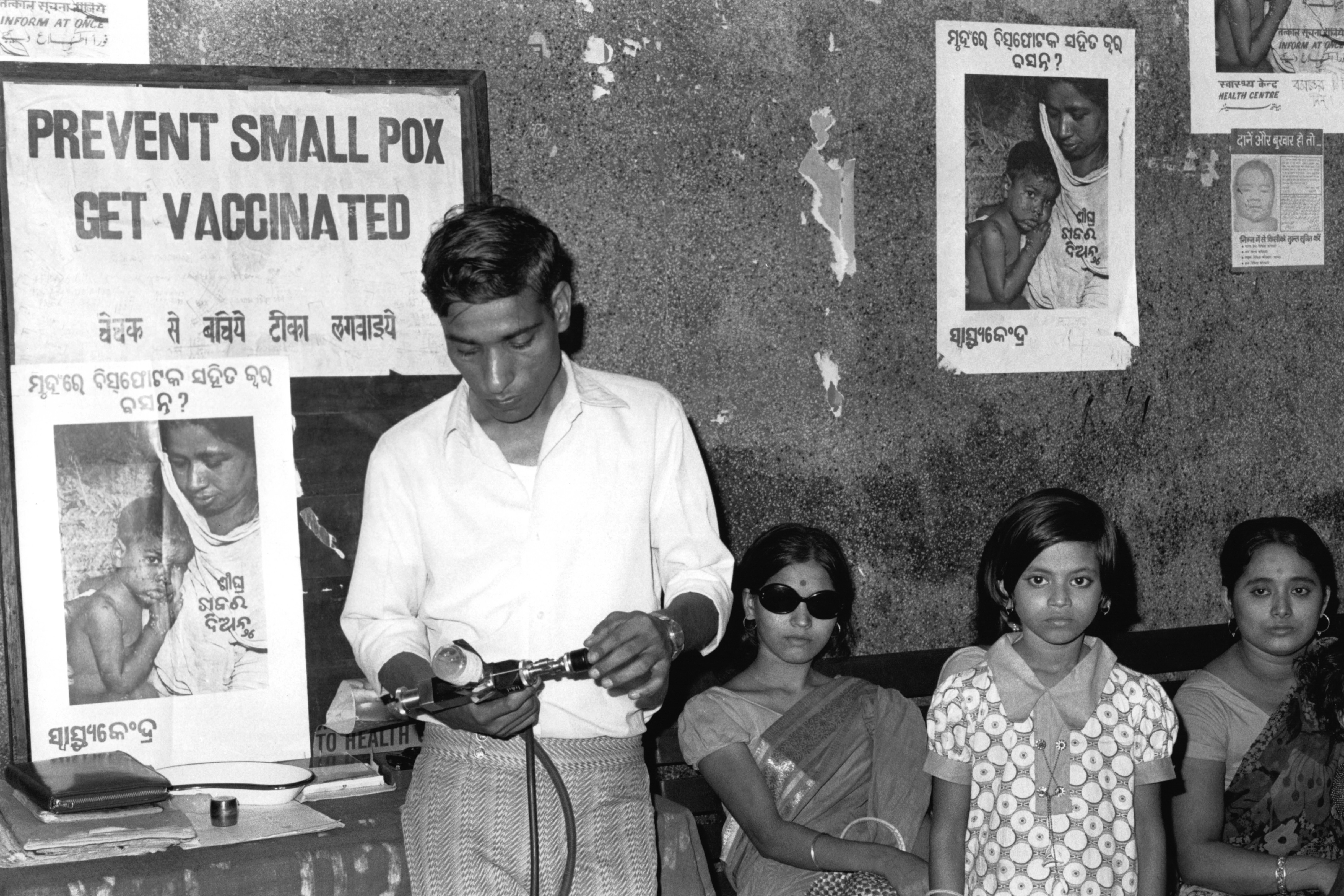 In this 1974 black-and-white film photograph, a health worker stands beside two women and a young girl who are waiting to be vaccinated. He holds a jet injector – a gun-shaped medical instrument that uses compressed air to force liquid vaccine through the skin without a needle. Beside him and on the walls are posters that say, “Prevent Small Pox / Get Vaccinated,” and ones that show images of a mother holding a child with smallpox.