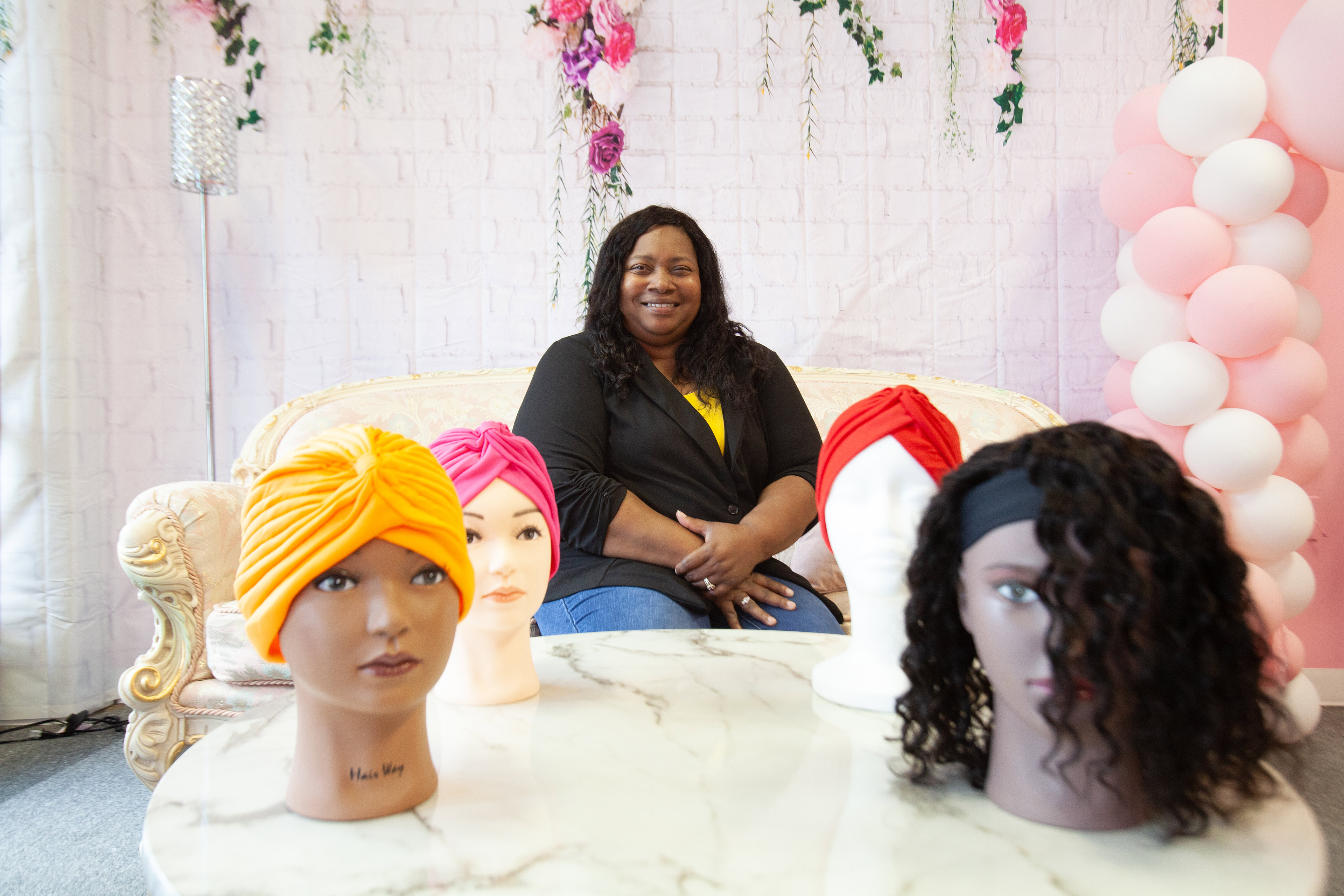 A photo of Kristie Fields sitting for a portrait indoors. A table with mannequin heads sporting hair wraps and wigs is in the foreground.