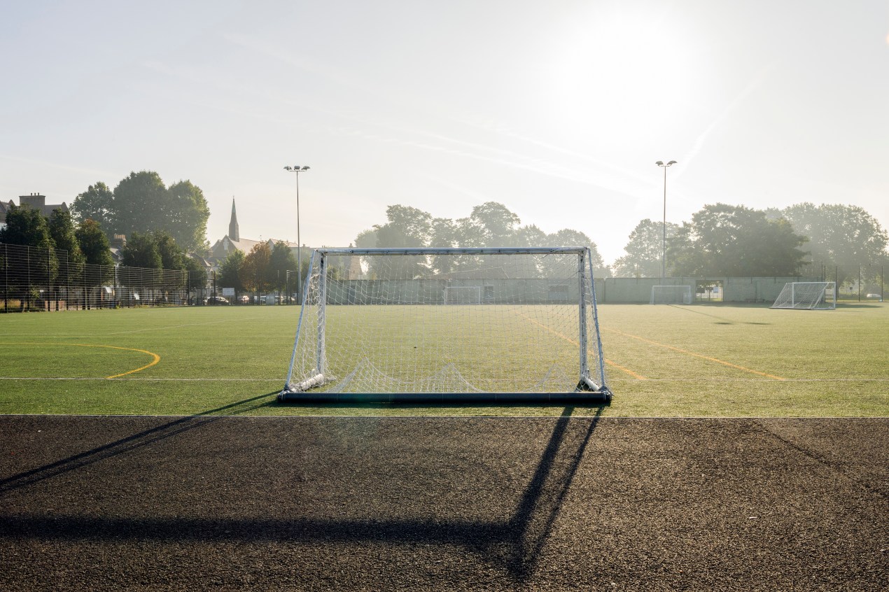 The photograph shows a sports field on a hot summer day. A soccer goal post, in the center of the image, casts a long shadow towards the viewer.
