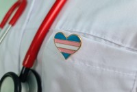 An up-close photograph of a heart-shaped pin in the colors of the transgender flag on a white doctor's coat. A red stethoscope is partially visible to the left of the pin.