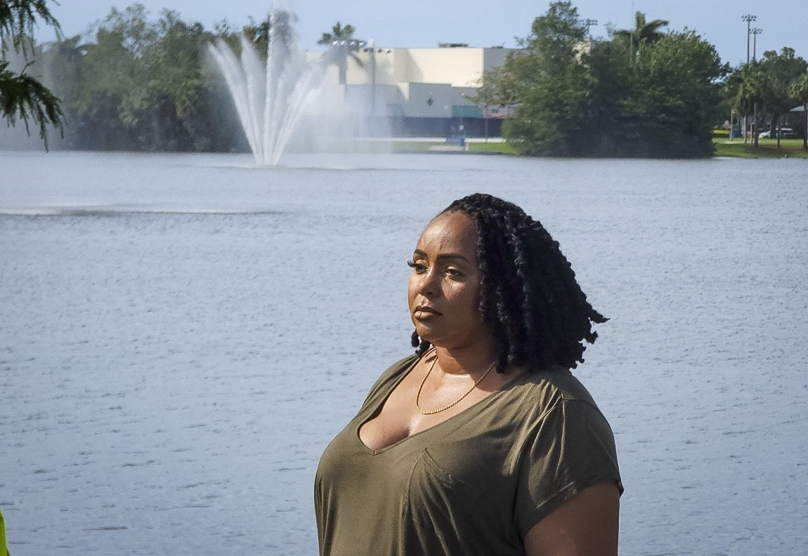 A portrait of Anya Cook standing in front of a body of water with a fountain. She looks off in to the distance with a contemplative expression.