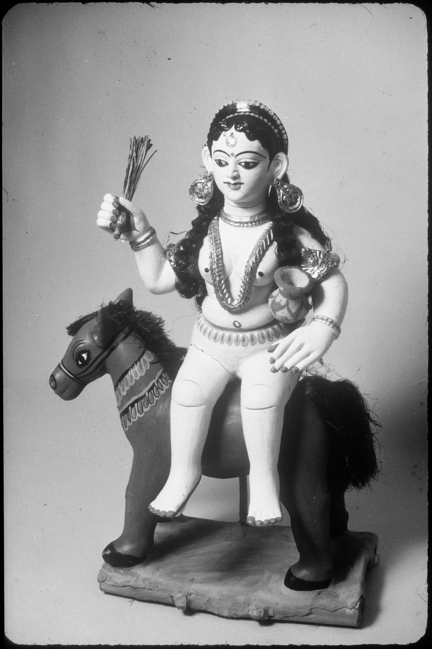 A black and white photo of a small statue of Shitala Mata, the Hindu goddess of smallpox. She is sitting on the back of a donkey. In her right hand, she holds up a small broom. In her left arm, she cradles a small pot which symbolically carries either the smallpox virus or clean water for healing from the virus.