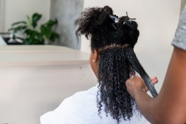 A photo of a black woman in a salon chair facing away from the camera. A stylist is applying cream to her naturally curly hair.