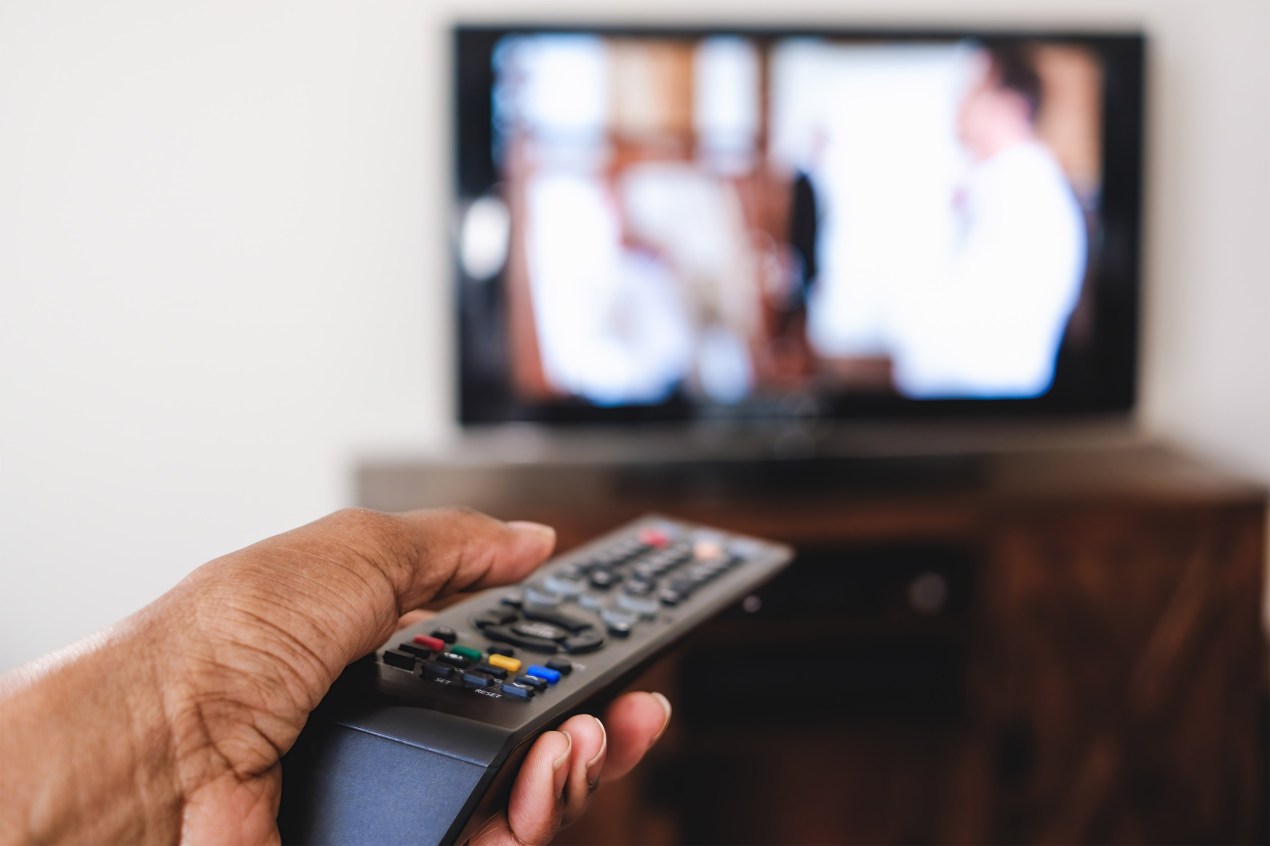 A photo of a hand holding a TV remote and pointing it at the screen.