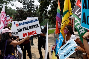 Two sides of a protest are seen outside. On the left are protesters against race-based affirmative action. One woman holds a sign that reads, "Stop discriminating on the basis of race." On the right are supporters of affirmative action. A woman on that side holds a sign that reads, "Diversity, opportunity, justice."