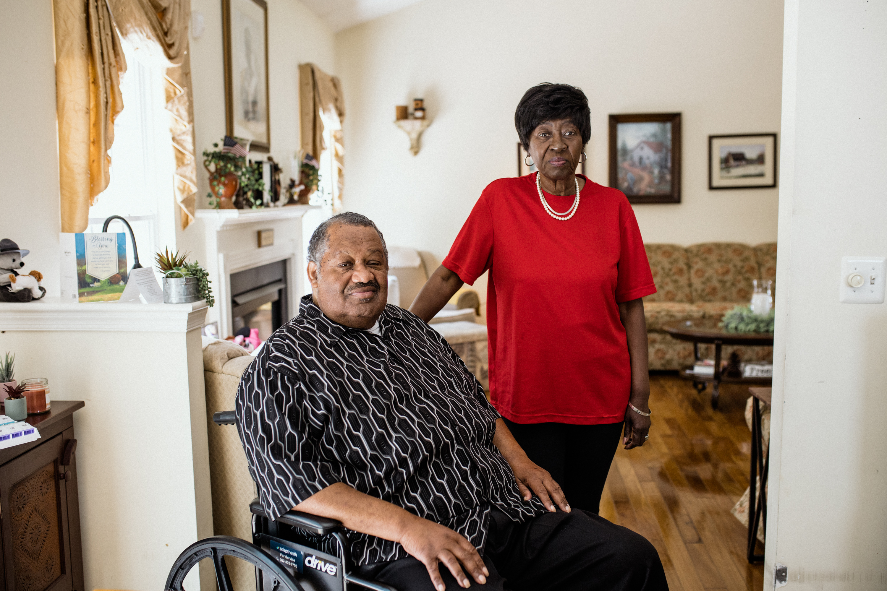 Thomas Greene is seated in a wheelchair with his wife standing beside him, her hand on his shoulder. Both subjects look towards the camera. They are in their home.
