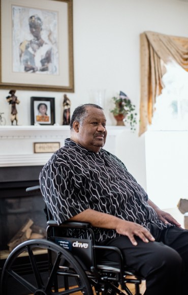 Thomas Greene is sitting in a wheelchair in his home, which is brightly lit by sunlight. He looks away from the camera.