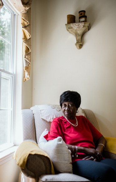 Bluizer Greene sits in a large, plush chair in a sun-lit corner of her home. Her hands are folded comfortably on her lap and she looks directly into the camera.