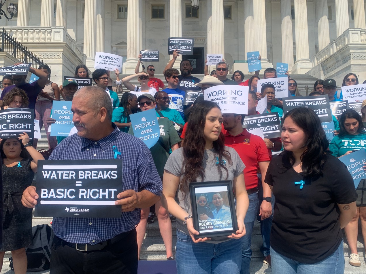 People of various ages and genders are standing in front of the U.S. Capitol building. Many hold signs that read, "Water Breaks = Basic Right". The female in the front of the line holds a framed photograph.