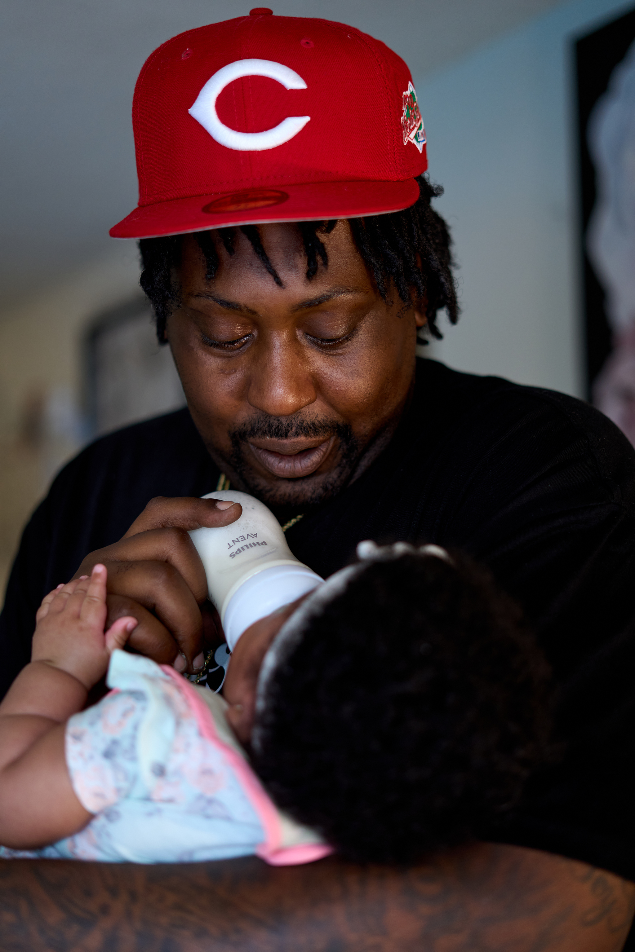 A Black man in a red sports cap cradles a newborn baby and feeds it a bottle