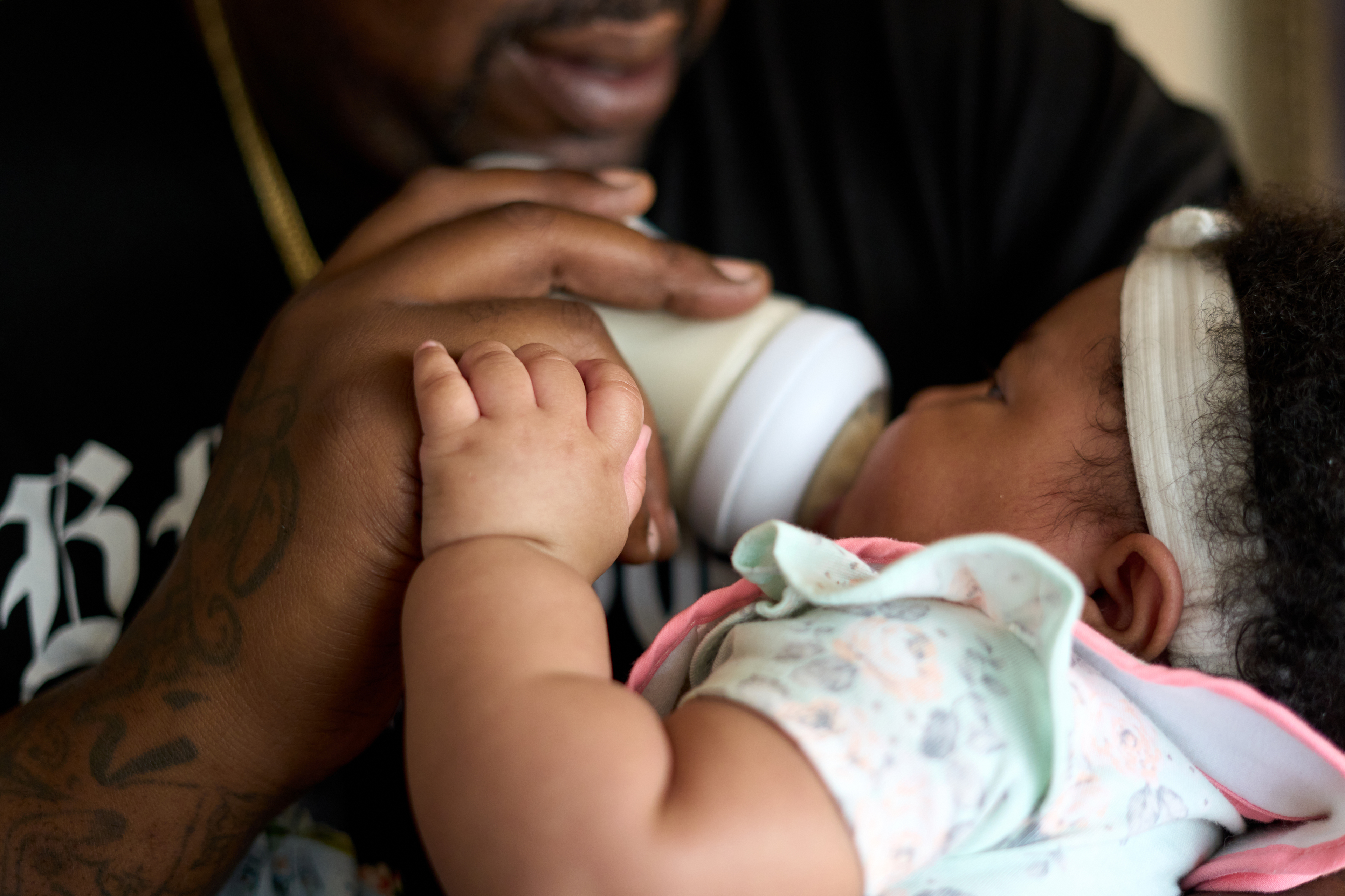 A father cradles his baby daughter and feeds her from a bottle. He is wearing a black t-shirt and she is wearing a white bow headband around her black curly hair.