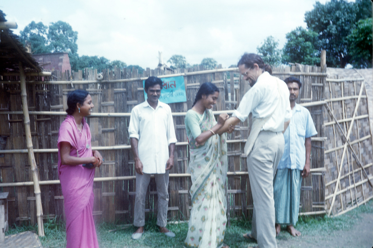 Steve Jones stands outside of a temporary smallpox isolation hospital in Bihar, India. The year is 1974 and the photograph was taken with color film. One woman and two men stand around Steve as a health worker vaccinates his lower left arm.